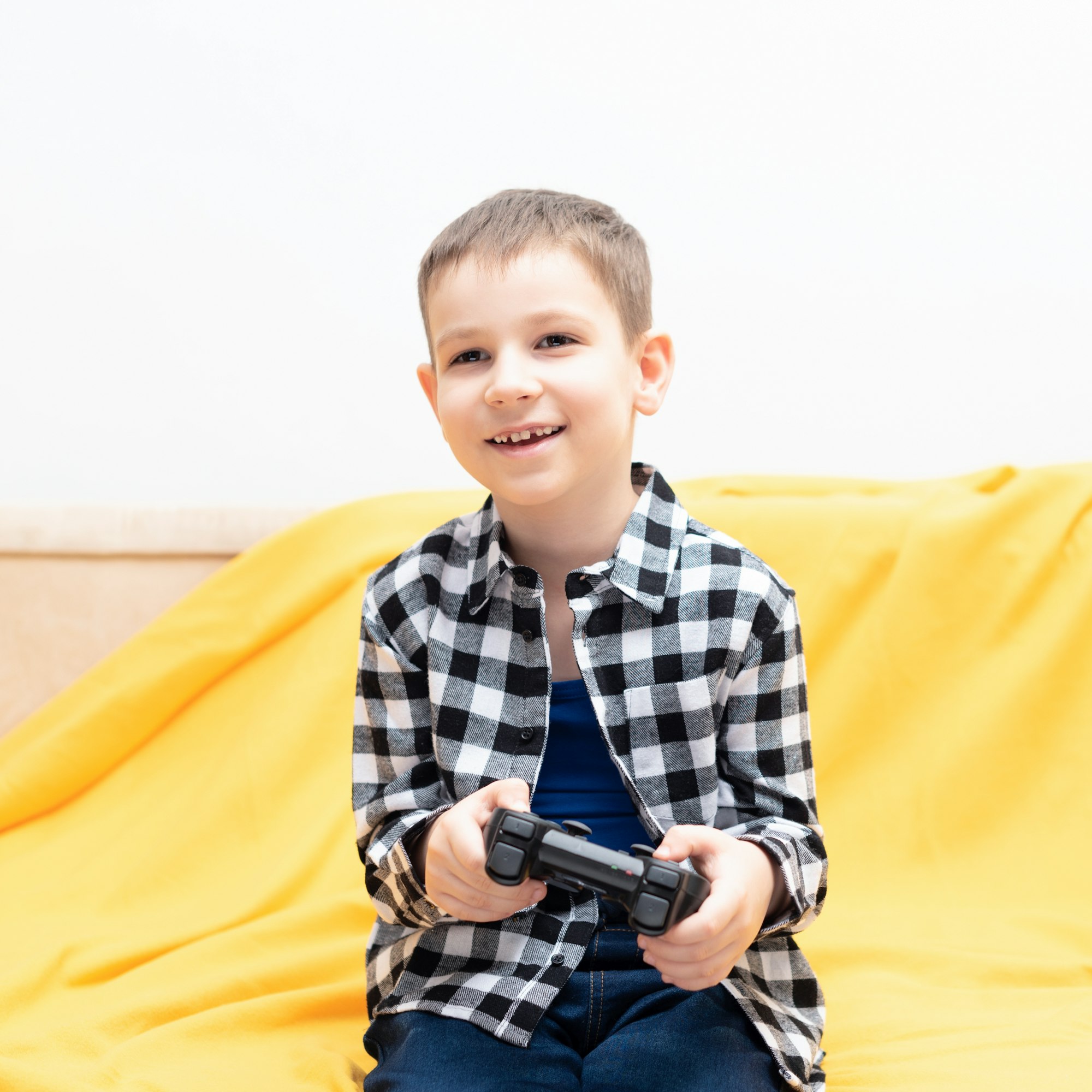 Happy child boy in checked shirt sitting on the couch with black joystick in his hands playing