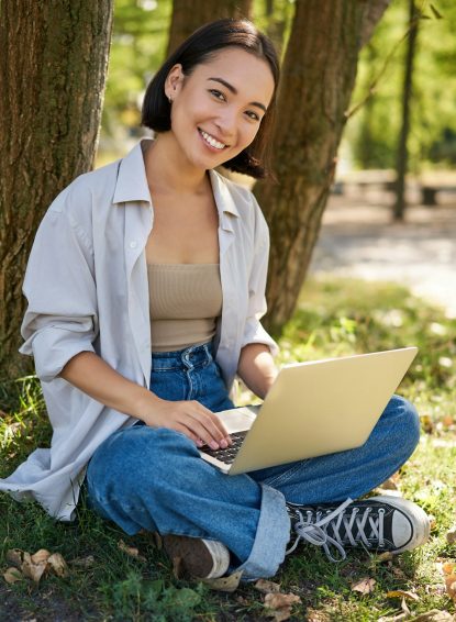 Beautiful asian girl sitting in park with laptop, working on remote, typing on keyboard, smiling at